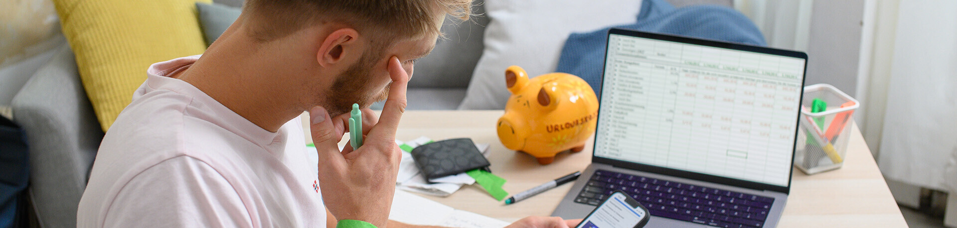 Mann sitzt vor seinem Laptop und seinem Handy und grübelt über Tabellen