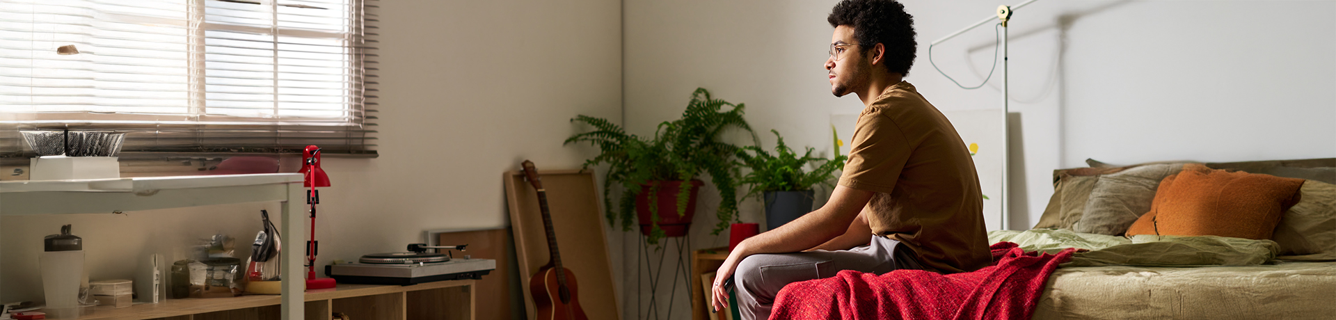 Junger Mann sitzt auf seinem Bett und schaut nachdenklich aus dem Fenster