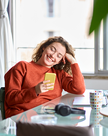 Junge Frau sitzt lächelnd an einem Tisch, hat den Kopf auf ihre Hand gestützt und schaut in ihr Handy