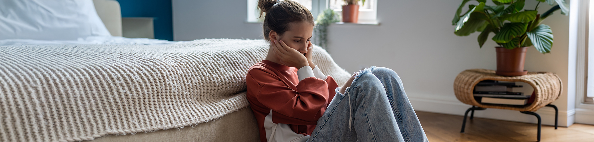 Junge Frau sitzt auf dem Boden vor ihrem Bett und hat verzweifelt ihren Kopf in ihre Hände gestützt