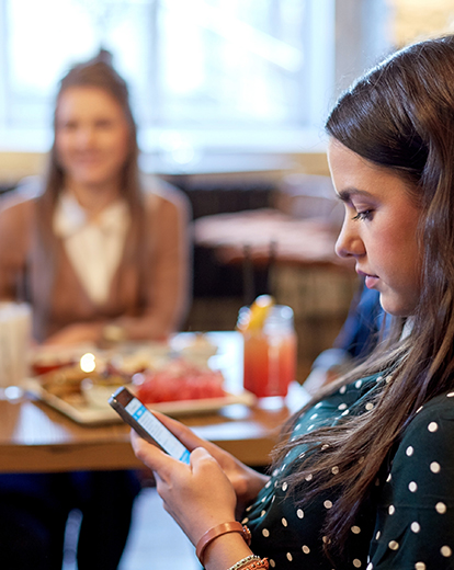 Junge Frau sitzt während dem Essen mit zwei Erwachsenen an ihrem Handy