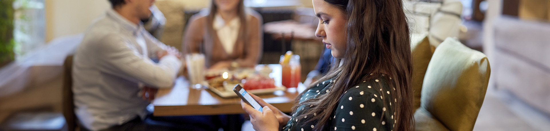 Junge Frau sitzt während dem Essen mit zwei Erwachsenen an ihrem Handy