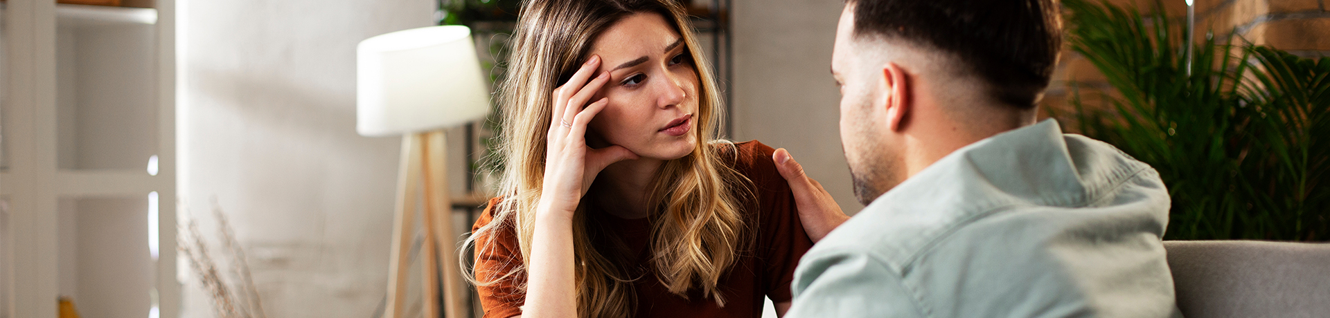 Junge Frau sitzt besorgt auf dem Sofa und unterhält sich mit Mann.