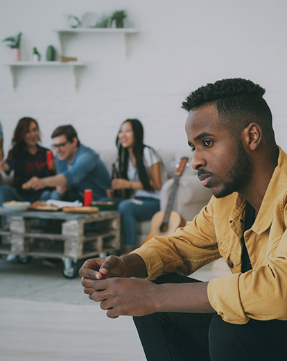 Junger Mann sitzt abgesondert von einer Gruppe nachdenklich auf einer Couch