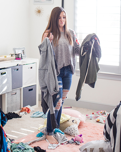 Junge Frau steht in der Mitte eines Zimmers mit Klamotten in beiden Händen