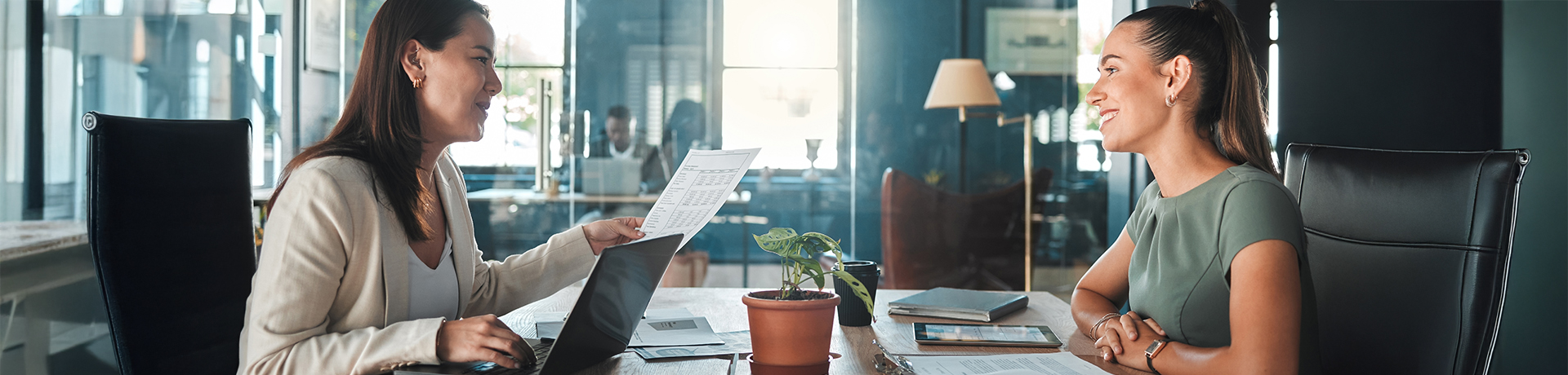 Junge Frau sitzt an einem Tisch und unterhält sich mit älterer Frau, die Unterlagen in der Hand hat