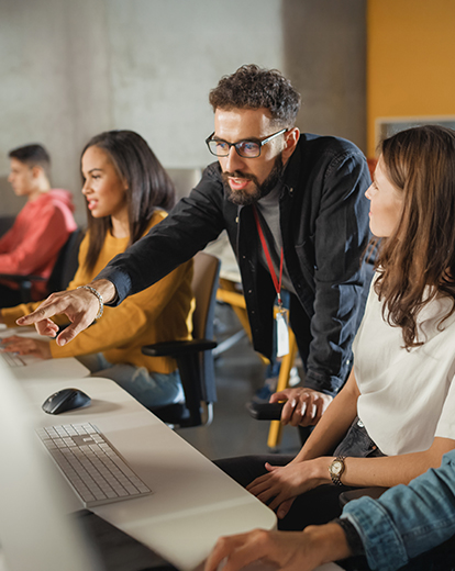 Junge Menschen sitzen an einem Tisch in einer Reihe, ein Mann beugt sich über den Computer