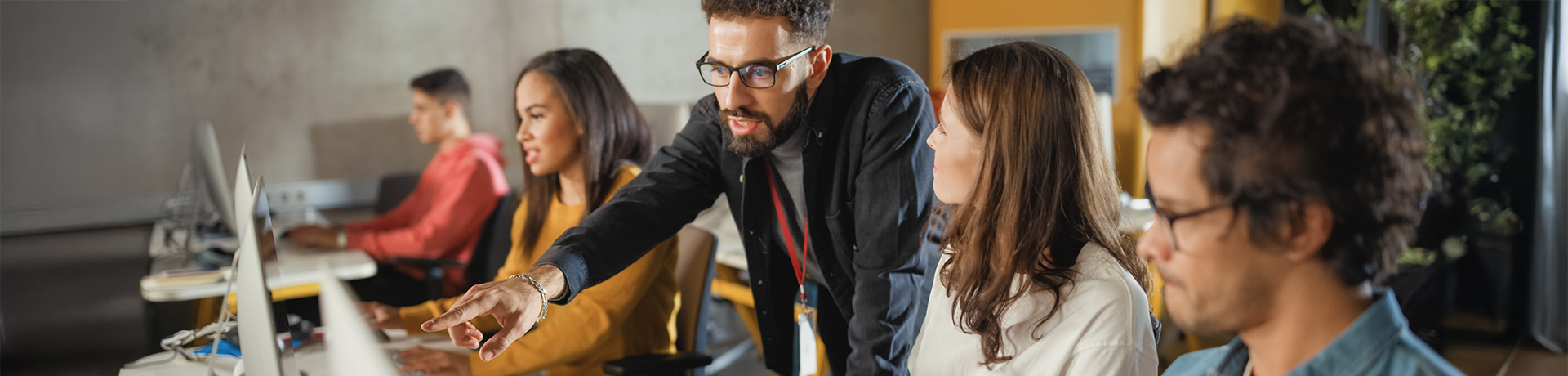 Junge Menschen sitzen an einem Tisch in einer Reihe, ein Mann beugt sich über den Computer