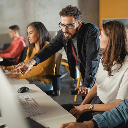 Junge Menschen sitzen an einem Tisch in einer Reihe, ein Mann beugt sich über den Computer