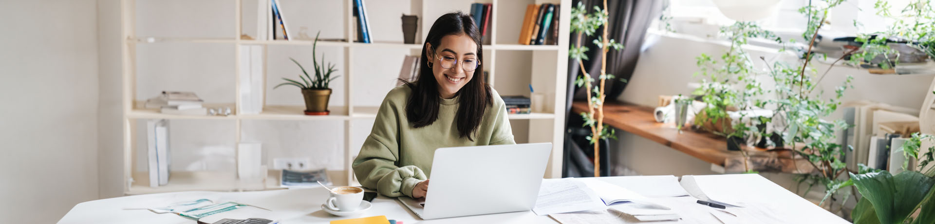 Junge Frau sitzt lächelnd vor ihrem Laptop an ihrem Arbeitsplatz
