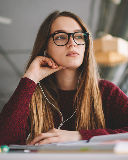 Junge Frau mit Kopfhörern sitzt an einem Tisch und schaut nachdenklich aus dem Fenster