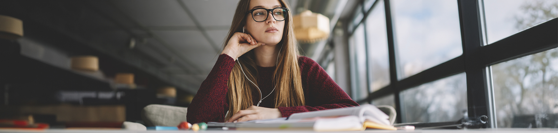 Junge Frau mit Kopfhörern sitzt an einem Tisch und schaut nachdenklich aus dem Fenster