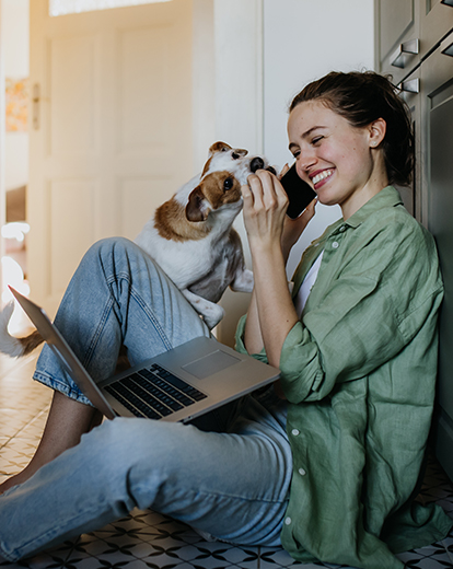 Junge Frau mit Laptop auf dem Boden sitzend wird von Hund abgeleckt