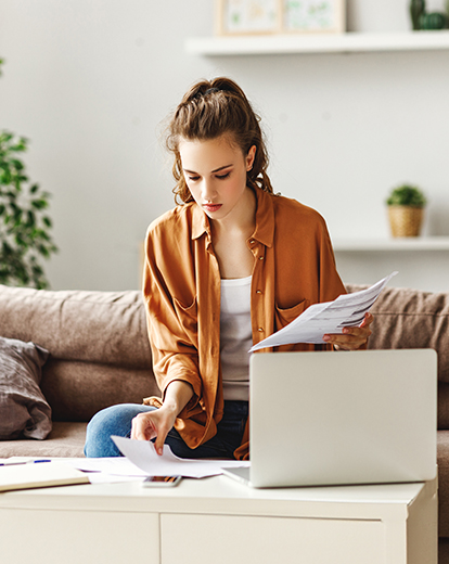 Junge Frau sitzt auf Sofa vor ihrem Laptop und schreibt auf Papier