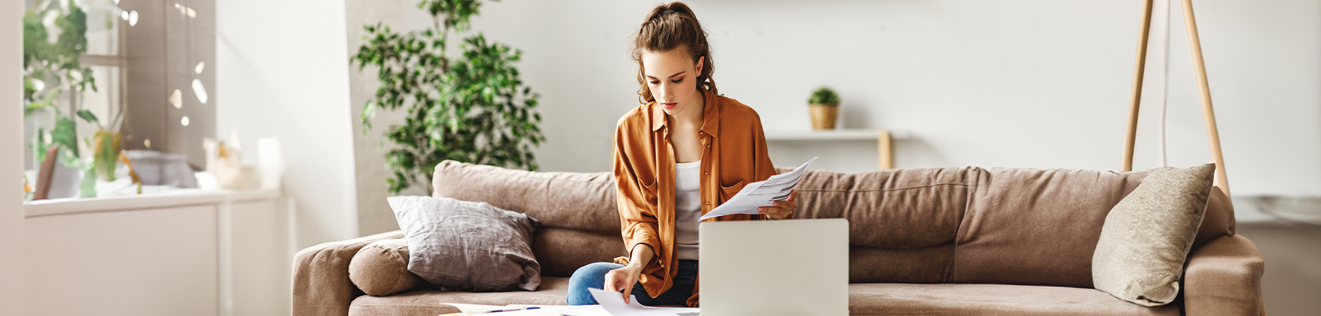 Junge Frau sitzt auf Sofa vor ihrem Laptop und schreibt auf Papier