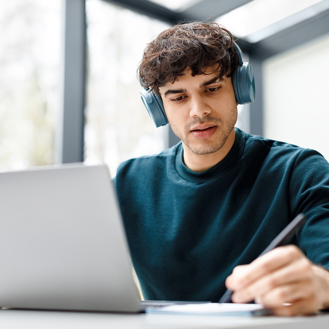 Ein junger Mann sitzt am Schreibtisch und macht sich Notizen während er auf seinen Laptop schaut.