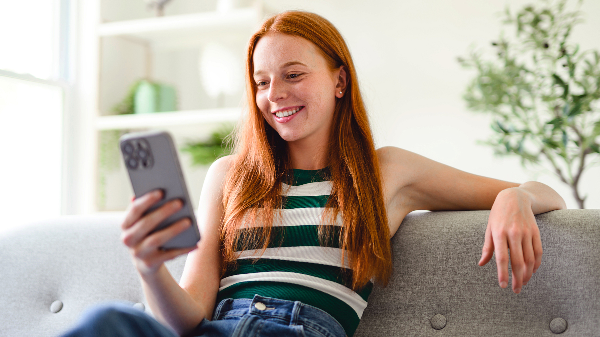 Eine Studentin sitzt entspannt auf dem Sofa und schaut lächelnd auf ihr Handy.