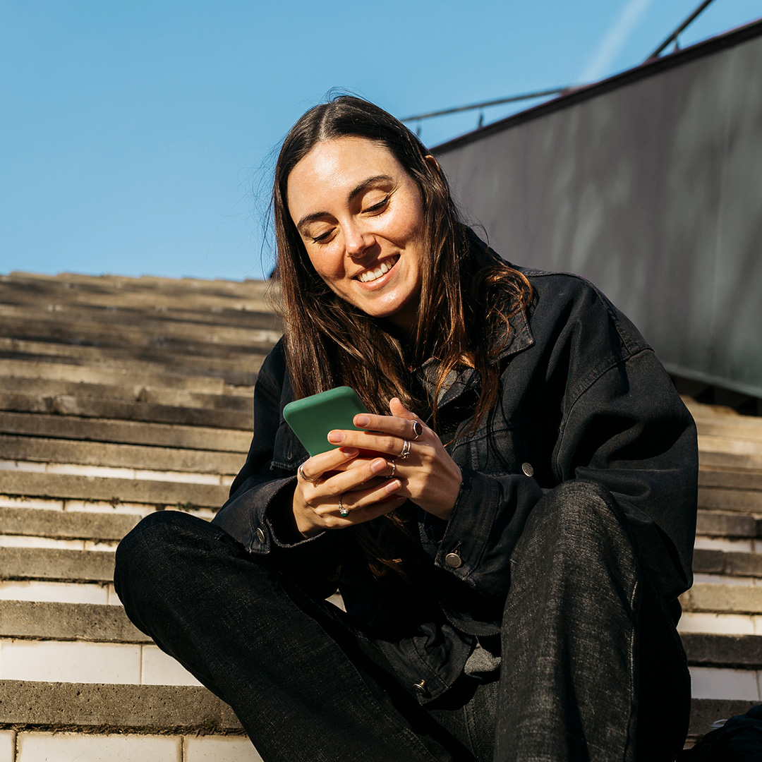 Eine Studentin sitzt auf Treppenstufen draußen und tippt gut gelaunt in ihr Handy.