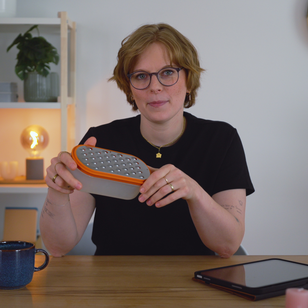 Dozentin Lisa Niendorf sitzt an einem Tisch und hält eine Käsereibe hoch.